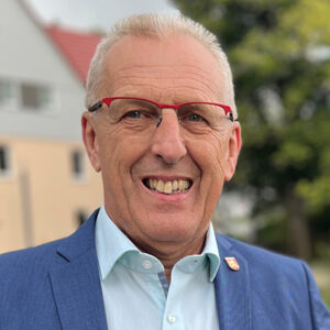 Älterer Mann mit Brille und blauem Anzug lächelt in die Kamera.