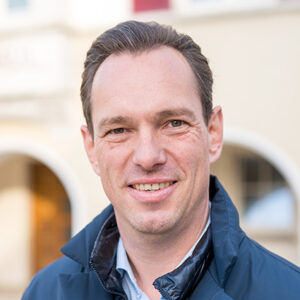 Ein Mann im Freien, trägt eine Jacke, lächelt in die Kamera.