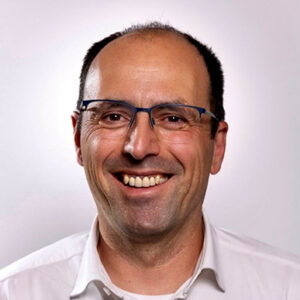 Mann mit Brille lächelt in die Kamera, trägt ein weißes Hemd.