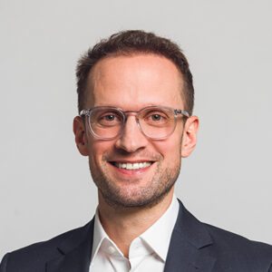 Mann mit Brille und Anzug lächelt vor grauem Hintergrund.