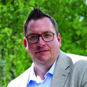 Mann mit Brille und hellem Sakko vor grünem, unscharfem Hintergrund.