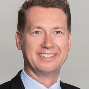 Ein lächelnder Mann in Anzug und Krawatte vor grauem Hintergrund.