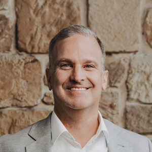 Eine Person in Anzug lächelt vor Steinmauer.