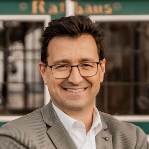 Mann mit Brille und Anzug lächelt, Hintergrund unscharf.