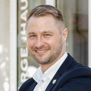 Ein lächelnder Mann in Anzug vor unscharfem Hintergrund.