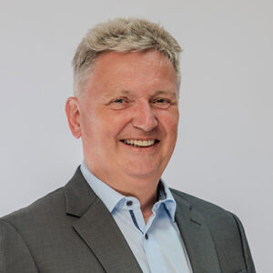 Ein lächelnder Mann in Anzug vor grauem Hintergrund.