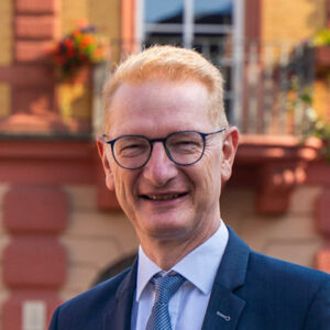 Ein lächelnder Mann mit Brille und Anzug vor einem Gebäude.