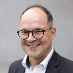 Mann mit Brille und Anzug lächelt vor grauem Hintergrund.