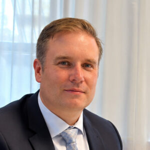 Eine Person in Anzug vor einem Fenster mit weißen Vorhängen.