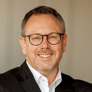 Ein lächelnder Mann mit Brille und Anzug vor neutralem Hintergrund.