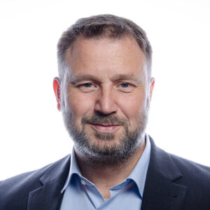 Ein Mann mit Bart, Hemd und Sakko lächelt in die Kamera vor hellem Hintergrund.