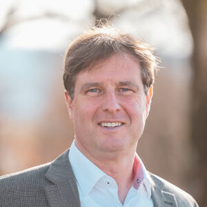 Ein Mann im Anzug steht im Freien und lächelt in die Kamera.