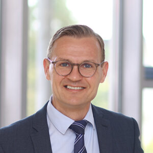 Mann in Anzug und Krawatte vor Glasfenster.