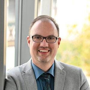 Mann mit Brille und Anzug lächelt vor einem Fenster.