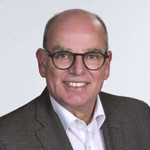 Mann mit Brille, Glatze, grauem Anzug und weißem Hemd lächelnd vor neutralem Hintergrund.
