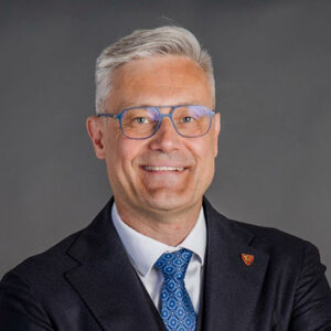 Person mit Brille und Anzug lächelt, grauer Hintergrund.