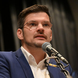 Mann mit Brille spricht in Mikrofon, trägt Anzug und Hemd.