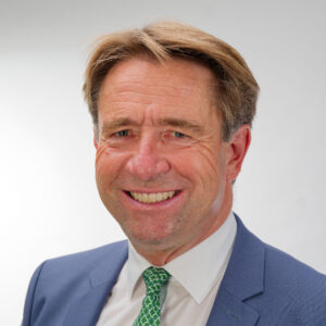 Ein lächelnder Mann in Anzug und Krawatte vor grauem Hintergrund.