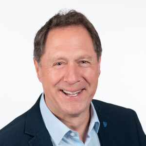 Mann in Anzug lächelt in die Kamera vor weißem Hintergrund.