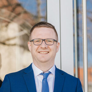 Mann in blauem Anzug und Krawatte, trägt Brille, lächelt in die Kamera.