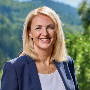 Eine lächelnde Frau im Anzug vor unscharfem, grünem Hintergrund.