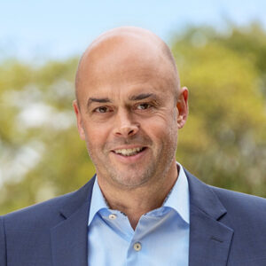 Ein Mann in Anzug lächelt in die Kamera, unscharfer Hintergrund.