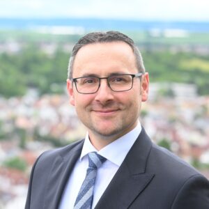 Mann mit Brille und Anzug vor unscharfem Hintergrund im Freien.