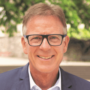 Ein lächelnder Mann mit Brille, im Anzug, vor unscharfem Hintergrund.