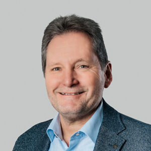 Ein lächelnder Mann in Anzug und Hemd vor grauem Hintergrund.