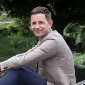 Ein Mann in beigem Sakko sitzt draußen vor grünen Bäumen.