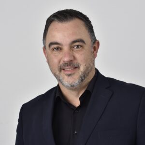 Mann mit Bart in dunklem Anzug vor grauem Hintergrund.