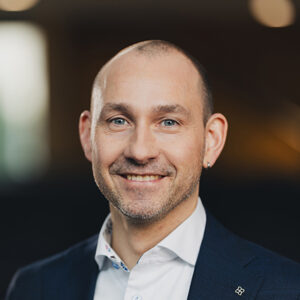 Lächelnder Mann in Anzug mit Ohrring, unscharfer Hintergrund.