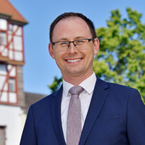 Mann im Anzug lächelt, Fachwerkhaus und Bäume im Hintergrund.