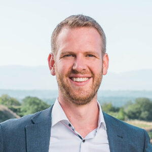 Ein Mann in Anzug lächelt im Freien. Hintergrund mit Bäumen und Hügeln.