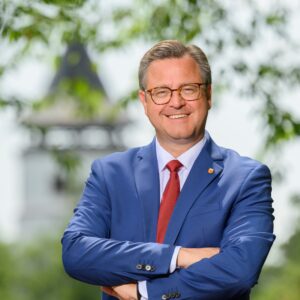 Mann in blauem Anzug mit roter Krawatte, lächelnd, im Freien, unscharfer Turm im Hintergrund.