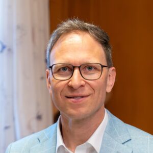 Ein Mann mit Brille und hellem Sakko vor einem Vorhang.