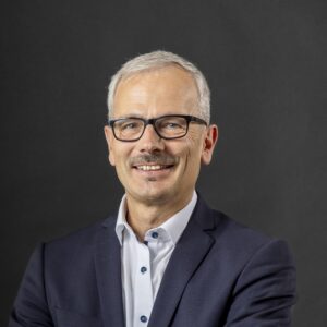 Eine lächelnde Person mit Brille und Anzug vor grauem Hintergrund.
