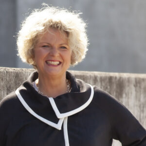 Eine lächelnde Person mit lockigem Haar vor einer Betonwand.