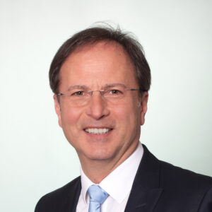 Mann mit Brille und Anzug lächelt vor hellem Hintergrund.