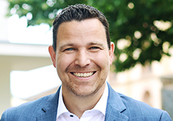 Ein lächelnder Mann in Anzug vor unscharfem Hintergrund.