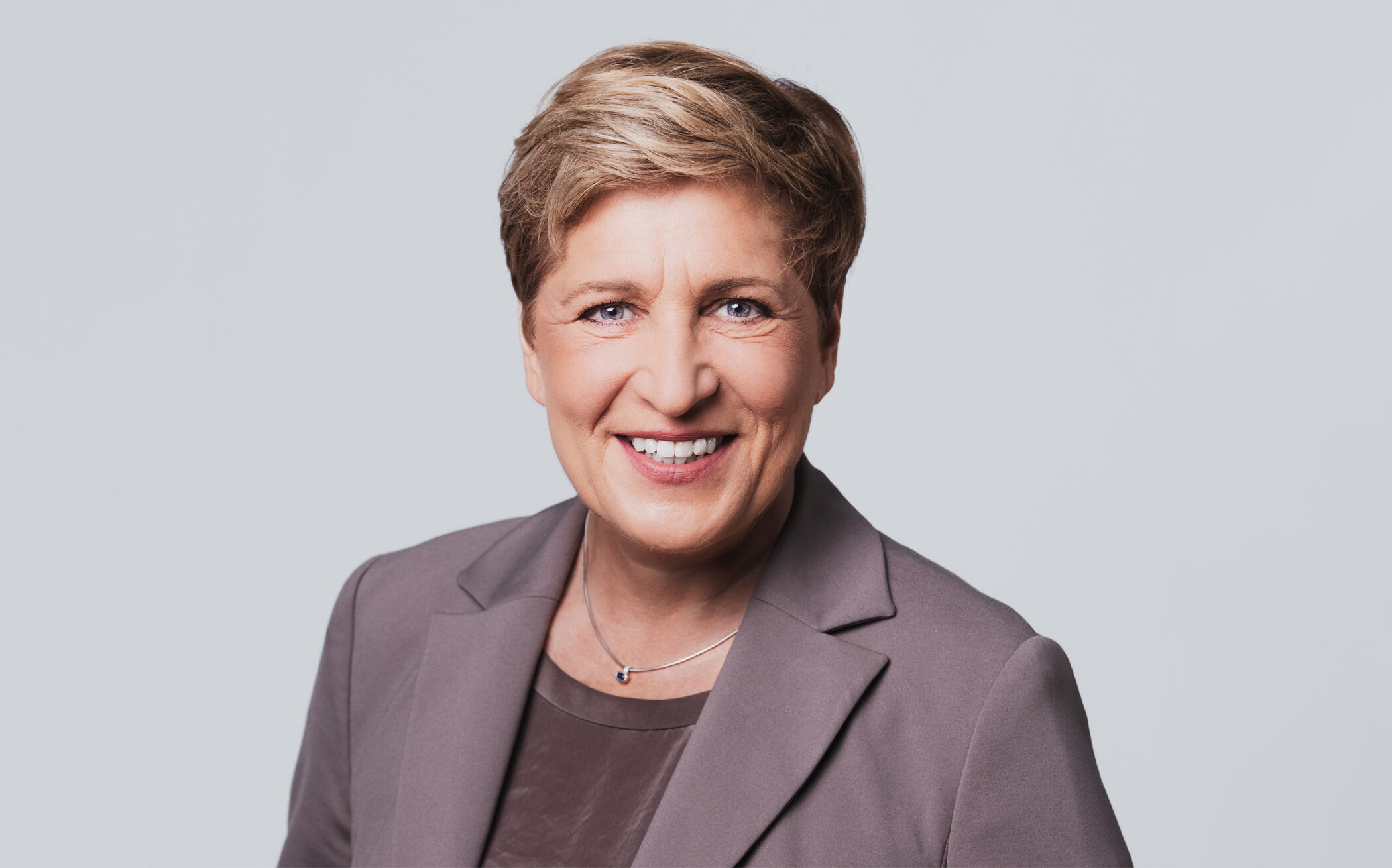 Eine Person in einem grauen Blazer lächelt vor neutralem Hintergrund.