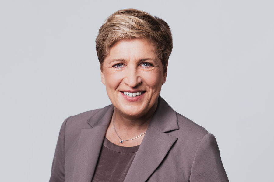 Eine Person in einem grauen Blazer lächelt vor neutralem Hintergrund.