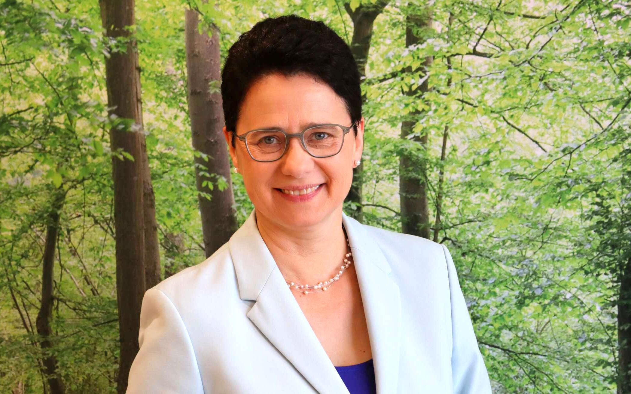 Eine Person mit Brille vor grünem Waldhintergrund, trägt ein helles Sakko.