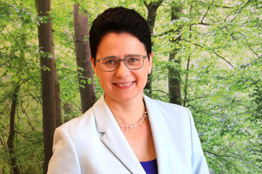 Eine Person mit Brille vor grünem Waldhintergrund, trägt ein helles Sakko.