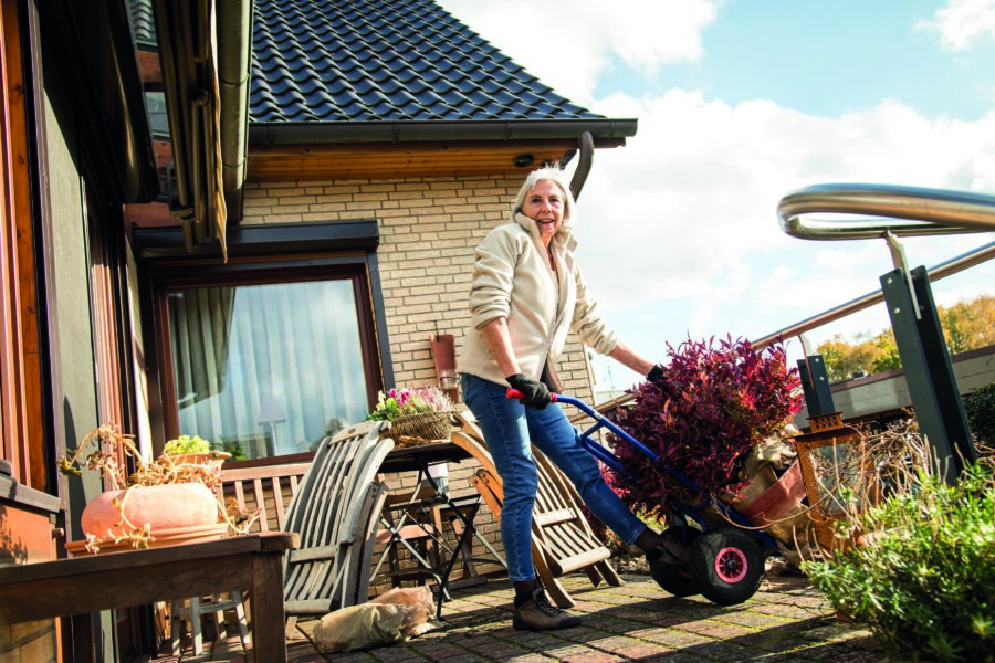 Eine ältere Dame stellt in einem Garten einen Pflanzentopf mit einer Sackkarre um