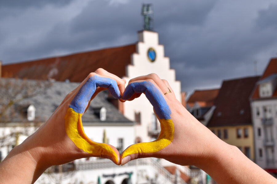 Herz mit Händen, welches mit Ukraine Farben ausgemalt ist, vor einem Rathausplatz