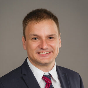 Ein Mann in Anzug und Krawatte lächelt vor grauem Hintergrund.