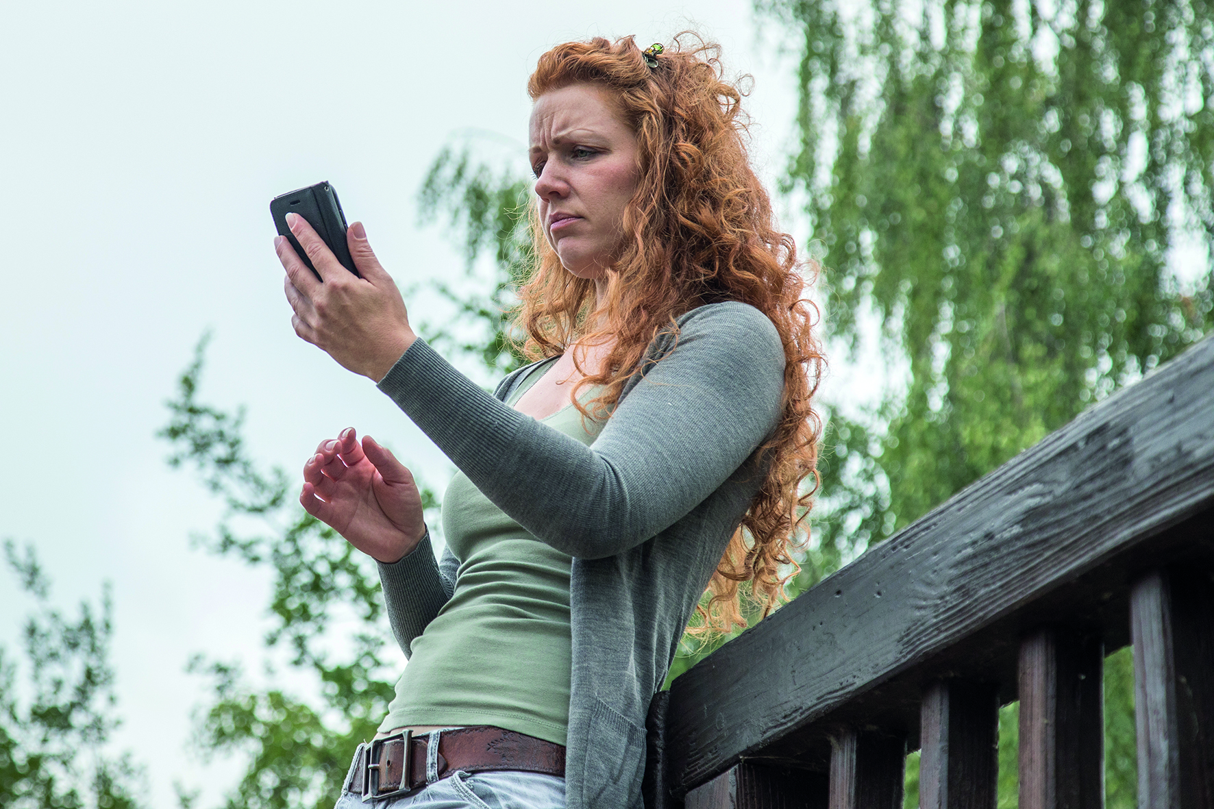 Eine junge Frau steht in einem Park auf einer Brücke und schaut auf ihr Smartphone