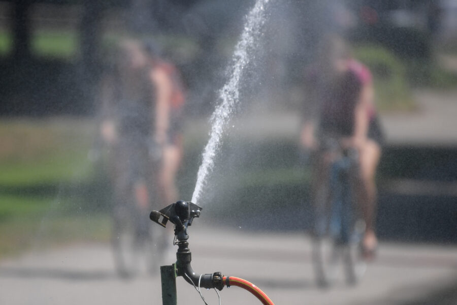 Junge Menschen fahren auf Fahrrädern an einem Wassersprenger vorbei.