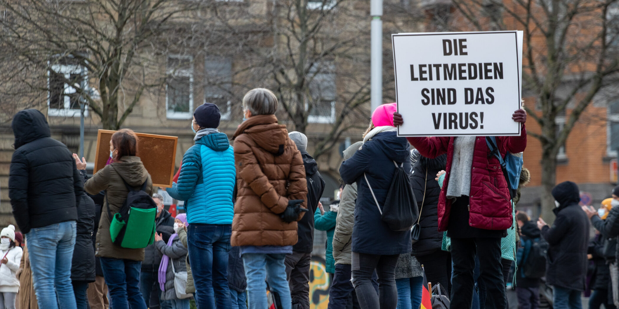 Frau mit Mundschutz hält ein Schild: Die Leidmedien sind das Virus!, dahinter Demo-Teilnehmer